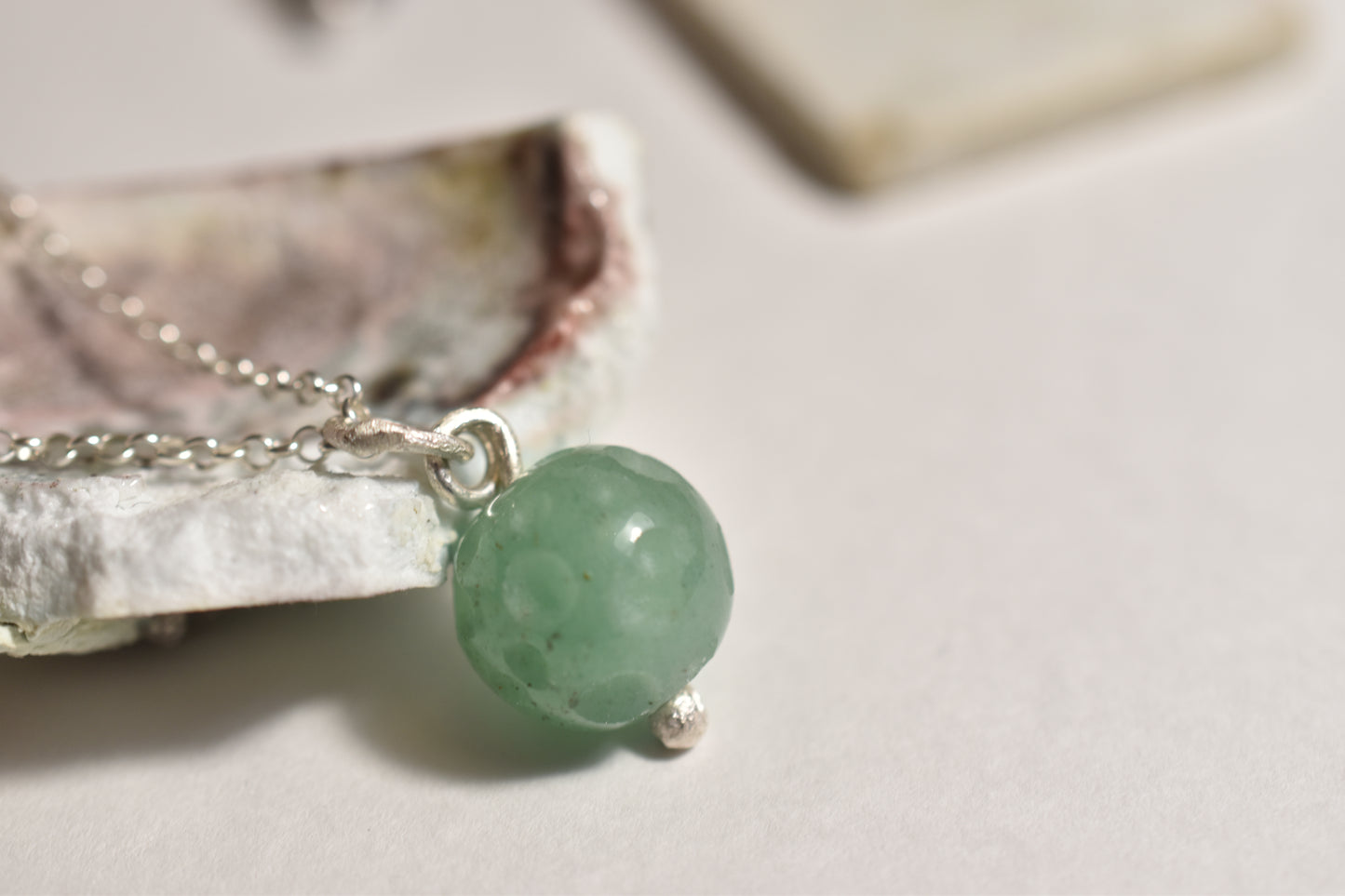 A carved round aventurine bead pendant with a silver setting, displayed on a white surface with a shell-like object in the background.